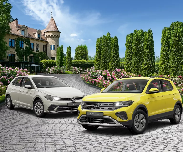 Zwei VW-Modelle in Gelb und Grau auf gepflastertem Hof vor einem Schloss mit blühenden Rosen und hohen Bäumen.