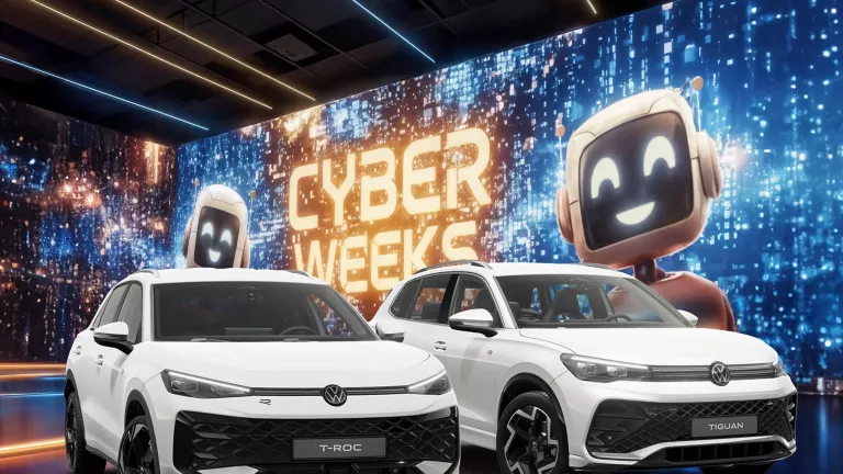 Zwei VW-Modelle vor einer großen digitalen Wand mit leuchtendem Schriftzug "CYBER WEEKS" und zwei lächelnden Roboter.
