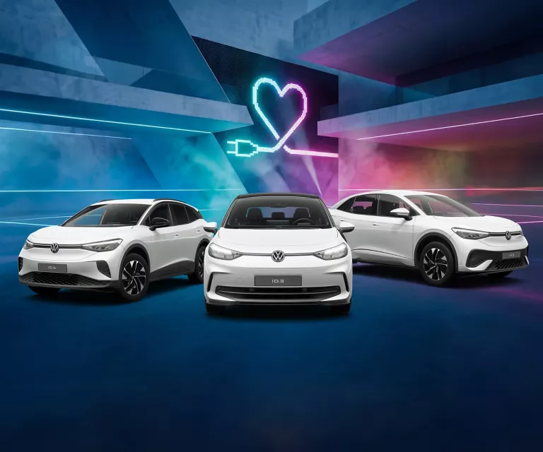 Drei VW Modelle vor Gebäude mit bunten LED's, im Hintergrund Herzsymbol mit Ladekabel-Stecker