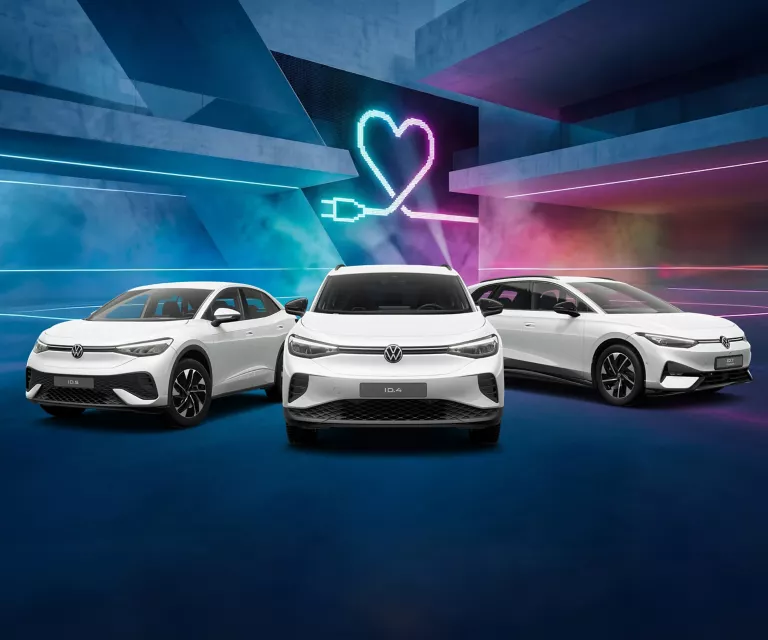 Drei VW Modelle vor Gebäude mit bunten LED's, im Hintergrund Herzsymbol mit Ladekabel-Stecker