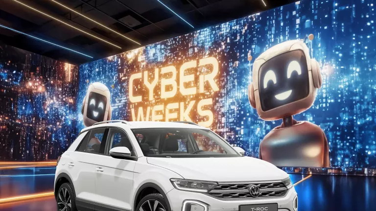 VW T-Roc R-Line in Weiß vor einer großen digitalen Wand mit leuchtendem Schriftzug "CYBER WEEKS" und zwei lächelnden Roboter.