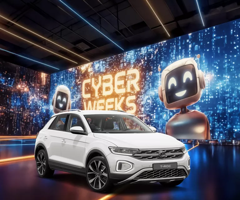 VW T-Roc R-Line in Weiß vor einer großen digitalen Wand mit leuchtendem Schriftzug "CYBER WEEKS" und zwei lächelnden Roboter.