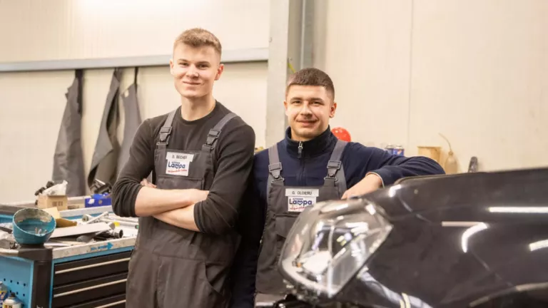 Zwei junge Männer stehen in grauer Latzhose hinter schwarzem Auto in Werkstatt