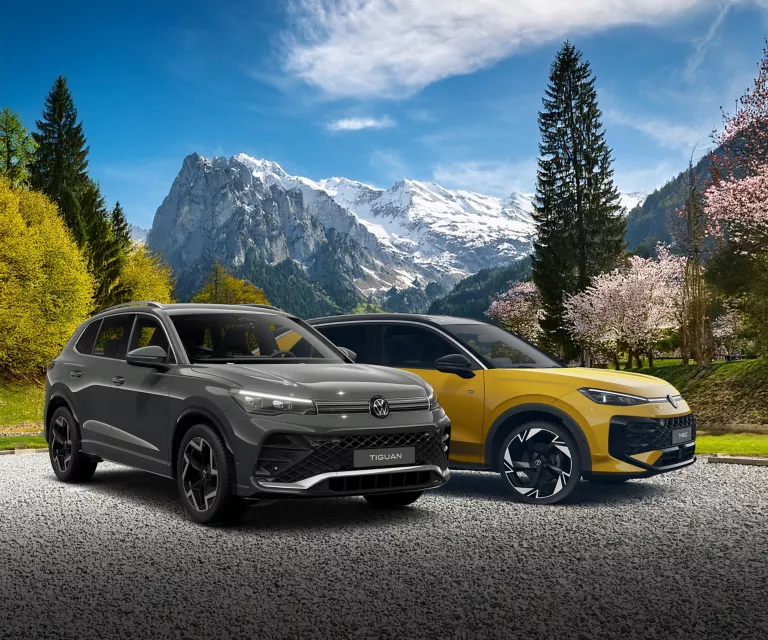 Ein VW Tiguan und ein V T-Roc parken auf einem Parkplatz. Im Hintergrund sieht man ein herrliches Berg Panorama.