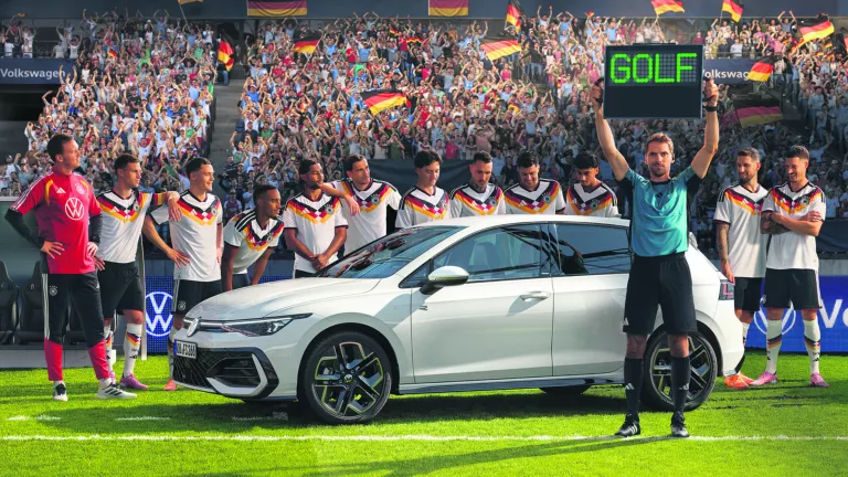 Ein weißer Golf R-Line steht mit der Herren Fußballnationalmannschaft auf einer Rasenfläche im Stadion. Der Schiedsrichter hält die Wechselanzeige mit dem Schriftzug „Golf“ in die Höhe. 