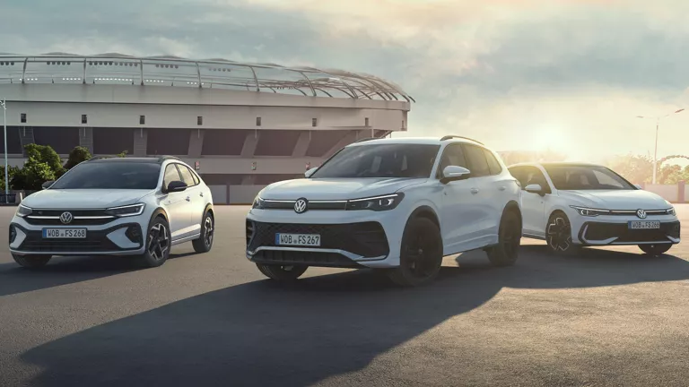 Ein weißer Tiguan R-Line, ein weißer Taigo und ein weißer Golf stehen auf dem Parkplatz eines Stadions im Gegenlicht. 