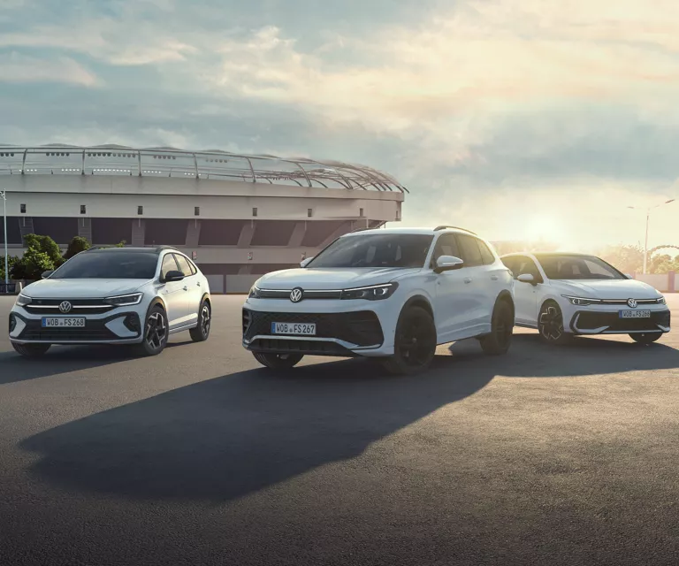 Ein weißer Tiguan R-Line, ein weißer Taigo und ein weißer Golf stehen auf dem Parkplatz eines Stadions im Gegenlicht. 