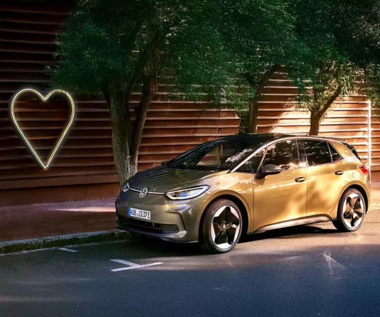 Ein Volkswagen ID.3 steht am Straßenrand unter grünen Bäumen vor einer modernen Holzfassade. Links im Bild leuchtet ein großes Herz-Symbol.