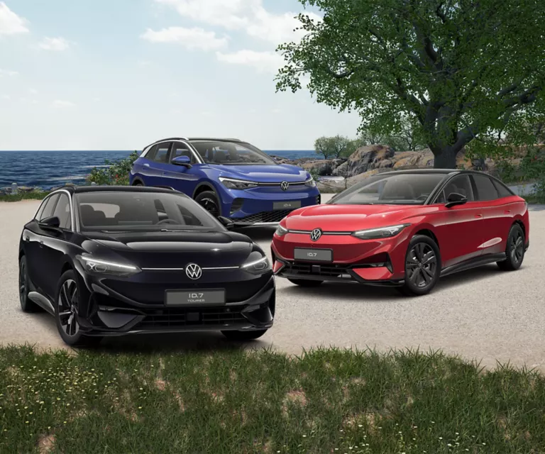 Drei VW Elektroautos stehen auf einem Kiesweg: ein schwarzer ID.7, ein blauer ID.4 und ein roter ID.7 Tourer. Im Hintergrund ist ein See und Bäume sichtbar.