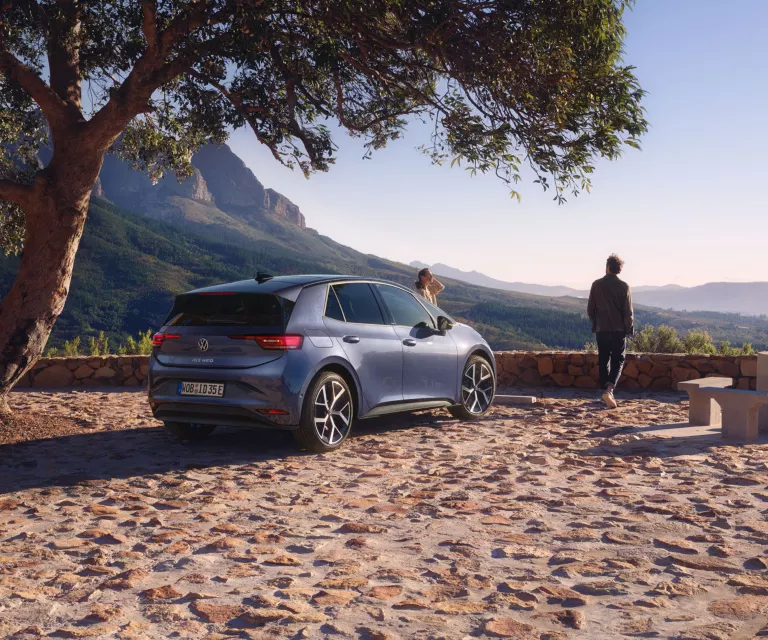 Auf einem Parkplatz in den Bergen parkt ein blauer VW ID.3 Neo, eine Frau und ein Mann genießen die Aussicht.