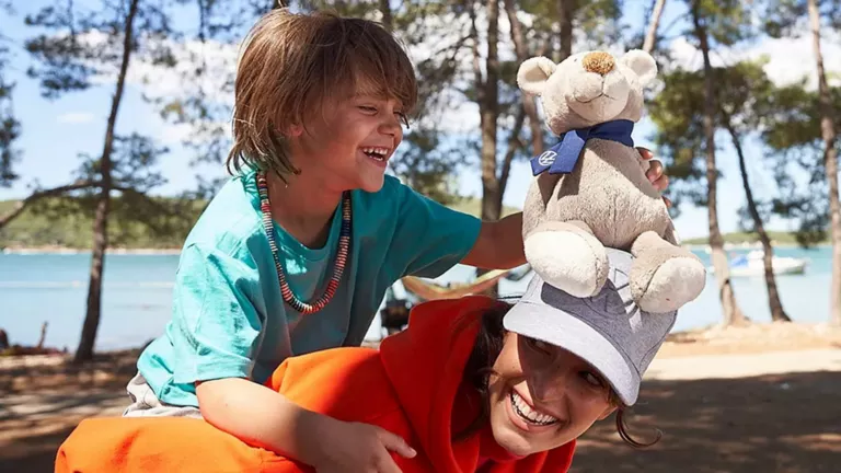 Kind lacht und sitzt auf den Schultern einer Frau mit grauer Kappe, die einen Plüschbären trägt, im Freien mit Bäumen und Wasser im Hintergrund