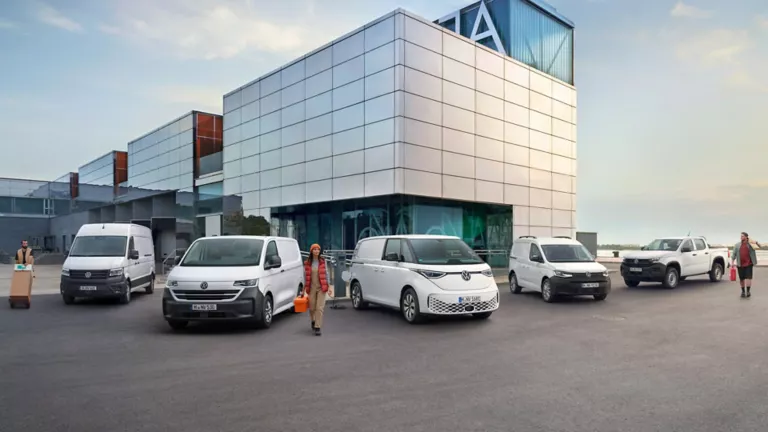 Sechs weiße Volkswagen Nutzfahrzeuge vor einem modernen Gebäude, zwei Personen tragen Pakete und Werkzeuge