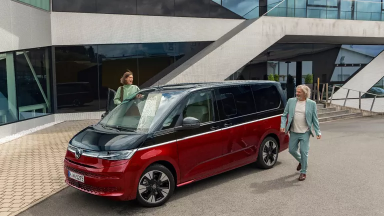 Zweifarbiger VW Multivan vor einem modernen Gebäude mit Sichtbeton und viel Glas in der Fassade.