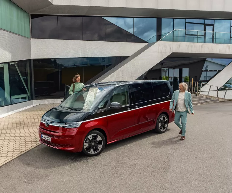 Zweifarbiger VW Multivan vor einem modernen Gebäude mit Sichtbeton und viel Glas in der Fassade.