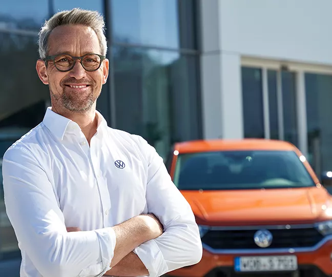 Ein Volkswagen Mitarbeiter steht mit verschränkten Armen vor einem VW Autohaus