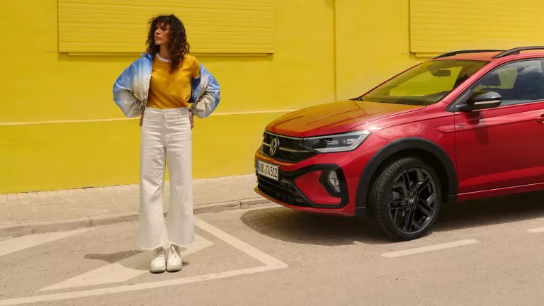 Roter VW Taigo parkt vor einem gelben Gebäude. Daneben eine junge Frau, die Hände in die Hüften gestemmt.