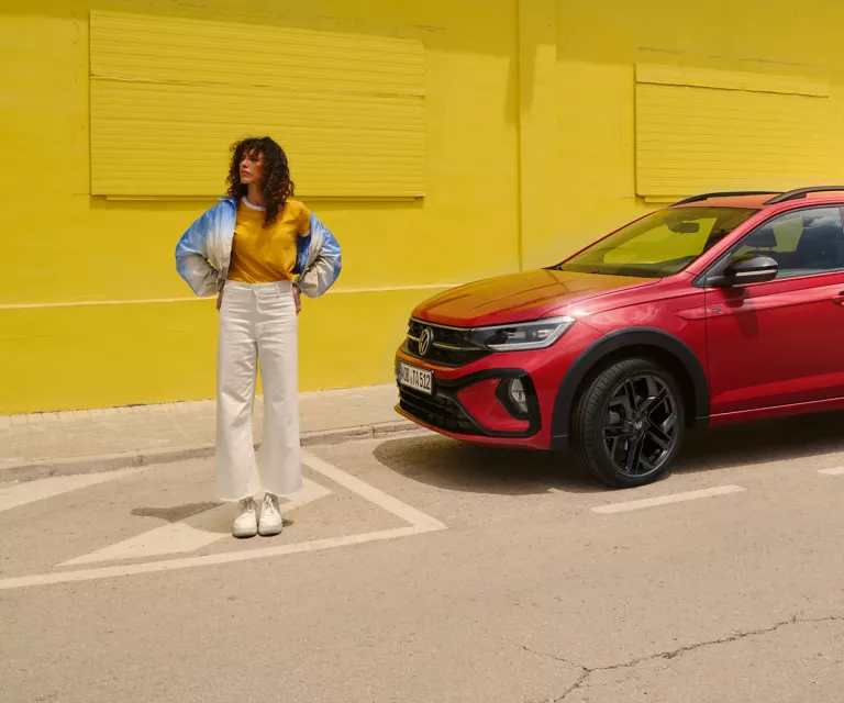 Roter VW Taigo parkt vor einem gelben Gebäude. Daneben eine junge Frau, die Hände in die Hüften gestemmt.