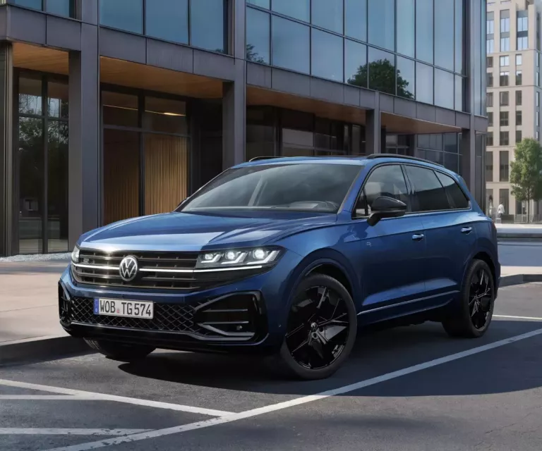 Ein dunkelblauer Touareg „FINAL EDITION“ parkt an einem Seitenstreifen vor einem futuristischen Gebäude. Im Hintergrund sind weitere Gebäude sowie grüne Bäume zu erkennen.