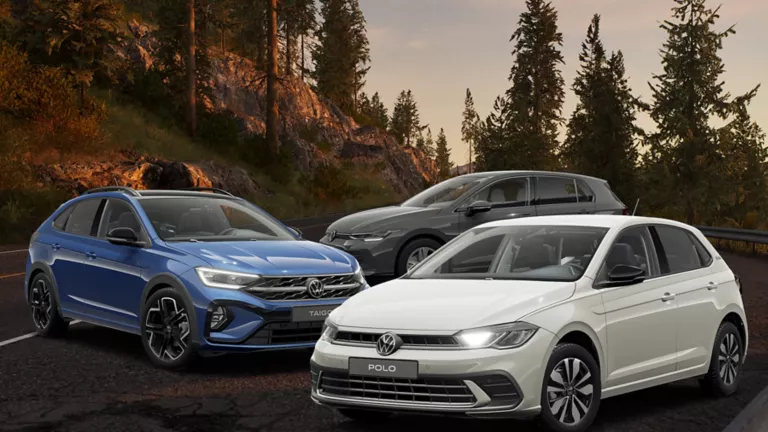 Ein blauer Volkswagen Taigo, ein grauer Polo und ein grauer Golf stehen auf einer Bergstraße bei Sonnenuntergang, umgeben von Kiefern.