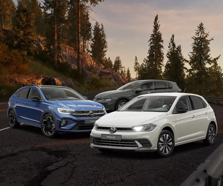 Ein blauer Volkswagen Taigo, ein grauer Polo und ein grauer Golf stehen auf einer Bergstraße bei Sonnenuntergang, umgeben von Kiefern.