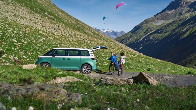 Ein VW ID.Buzz steht mitten in grünen Bergern. Im Hintergrund sind zwei Paraglider zu sehen.
