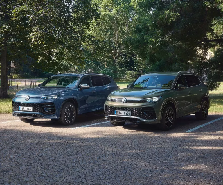Zwei geparkte VW SUV-Modelle in Blau und Grau auf gepflastertem Parkplatz vor Bäumen.