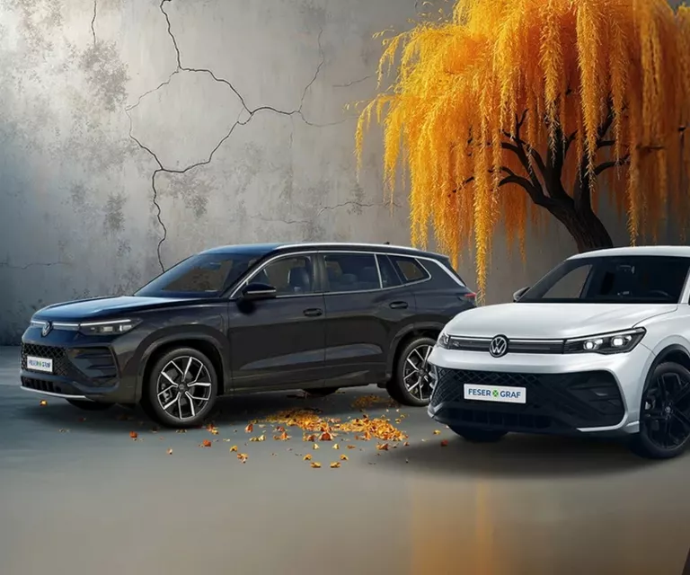 Zwei VW SUVs, Tiguan R-Line in Weiß und Tayron in Dunkelblau, vor einem modernen Hintergrund mit herbstlichem Baum.