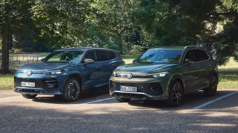 Blauer VW Tayron und grüner VW Tiguan stehen auf einem Parkplatz umgeben von Bäumen. 
