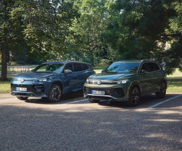 Blauer VW Tayron und grüner VW Tiguan stehen auf einem Parkplatz umgeben von Bäumen. 