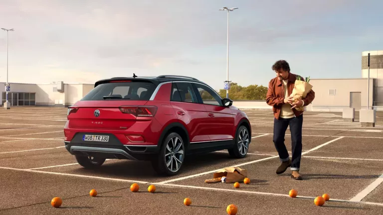 Bild zeigt einen T-Roc auf einem Parkdeck. Ein Mann mit einer Tüte Einkäufe steht daneben. Er hat Orangen verloren, die überall herumliegen.