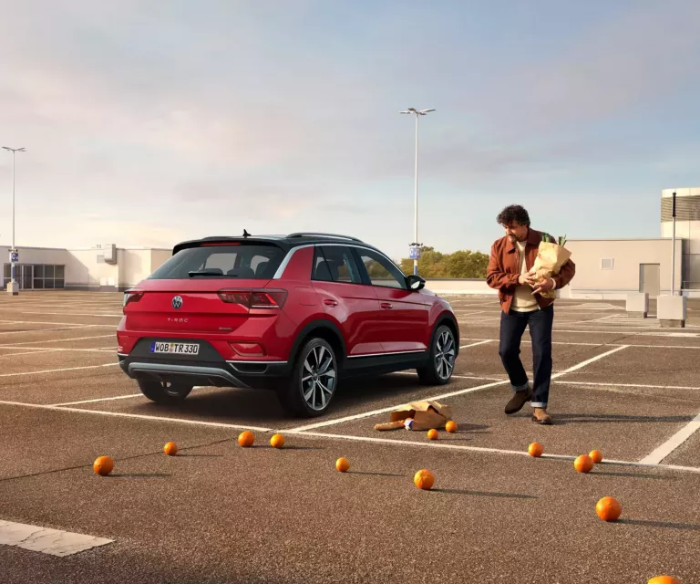 Bild zeigt einen T-Roc auf einem Parkdeck. Ein Mann mit einer Tüte Einkäufe steht daneben. Er hat Orangen verloren, die überall herumliegen.