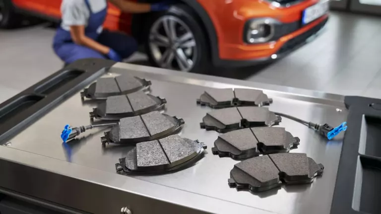 Acht Volkswagen Original Bremsbeläge auf einem Rollcontainer, im Hintergrund ein VW T-Cross – Bremsbeläge Auto