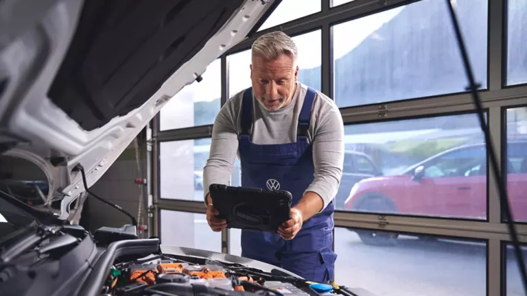Mechaniker in blauer Latzhose mit VW-Logo hält ein Tablet vor geöffneter Motorhaube eines Autos in einer Werkstatt mit großen Fenstern.