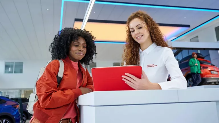 Kundin im roten Outfit spricht mit Mitarbeiterin in weißem Hemd mit Aufschrift 'Volkswagen Economy Service' an einem Empfangstresen, die auf ein rotes Tablet zeigt