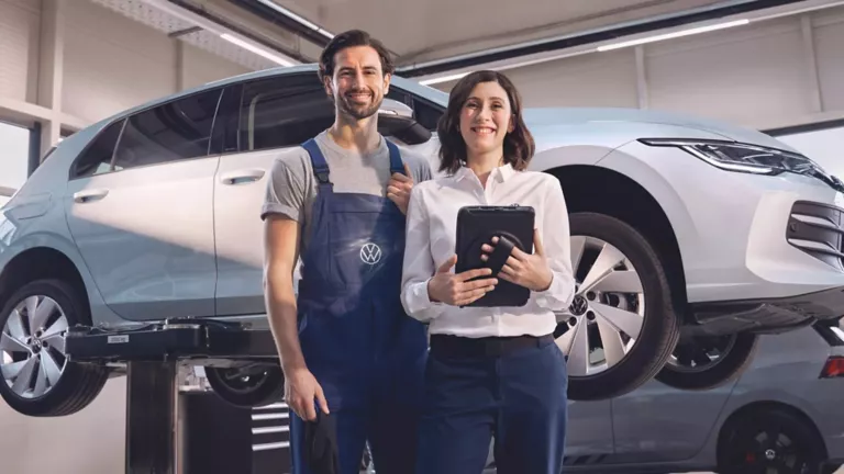 Zwei Volkswagen-Mitarbeitende, ein Mann in blauer Arbeitskleidung und eine Frau mit Tablet, stehen vor einem auf einer Hebebühne angehobenen weißen Volkswagen im Servicezentrum