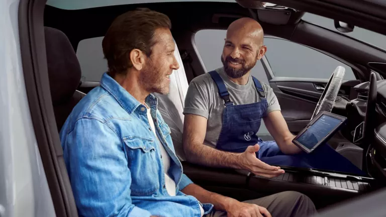 Ein Mechaniker im blauen Overall erklärt einem Kunden im blauen Jeanshemd im Auto mit Tablet in der Hand die Service-Terminplanung