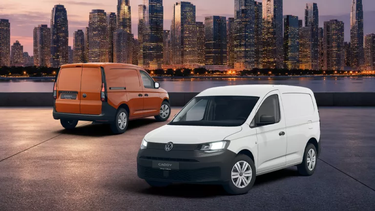 Zwei VW Caddy Cargo, ein weißer von vorne und ein orangeroter von hinten, vor einer beleuchteten Stadtsilhouette bei Dämmerung.  
