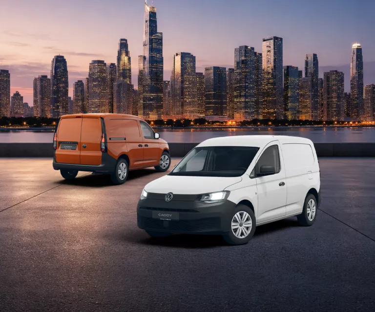 Zwei VW Caddy Cargo, ein weißer von vorne und ein orangeroter von hinten, vor einer beleuchteten Stadtsilhouette bei Dämmerung.  