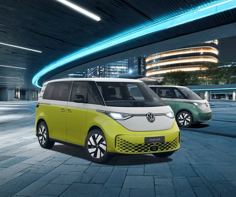 Zwei VW ID. Buzz Elektro-Vans in gelb-weiß und grün-weiß vor moderner, beleuchteter Architektur bei Nacht.