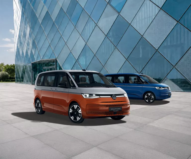 Zwei VW Multivan vor einem modernen Glasgebäude, ein orange-schwarzer und ein blau-schwarzer Van.