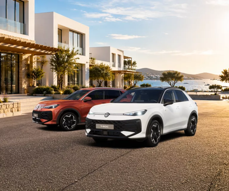 Zwei moderne VW T-Roc SUV, ein weißes und ein rotes, stehen vor einem modernen Haus mit Meerblick bei Sonnenuntergang.