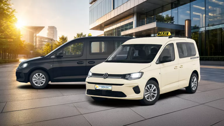 Vor einem modernen Glasgebäude: ein weißer VW Caddy mit Taxi-Dachzeichen, dahinter ein schwarzer VW Caddy.