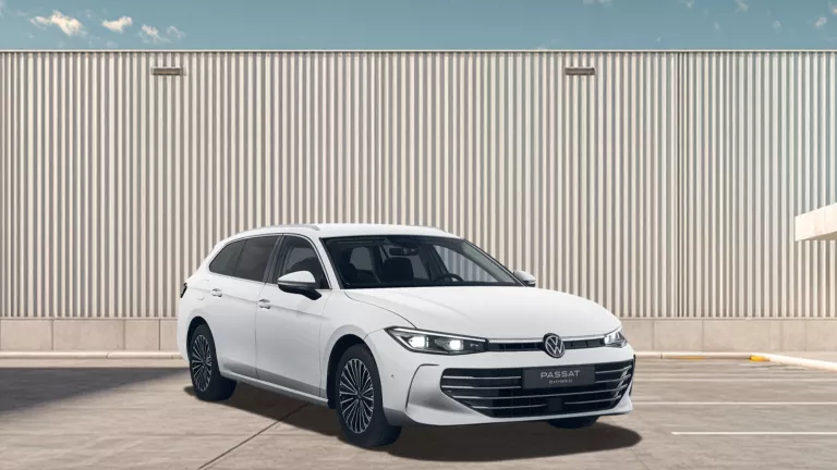 Weißer VW Passat steht auf einem leeren Parkplatz vor einer großen, hellen Wellblechwand unter blauem Himmel mit wenigen Wolken.