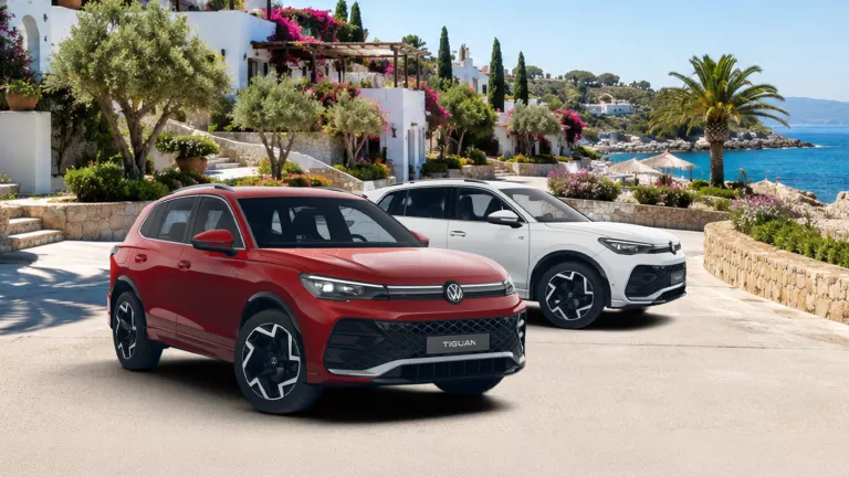 Zwei VW Tiguan, einer rot und einer weiß, stehen vor mediterraner Küstenlandschaft mit Meer und weißen Häusern.
