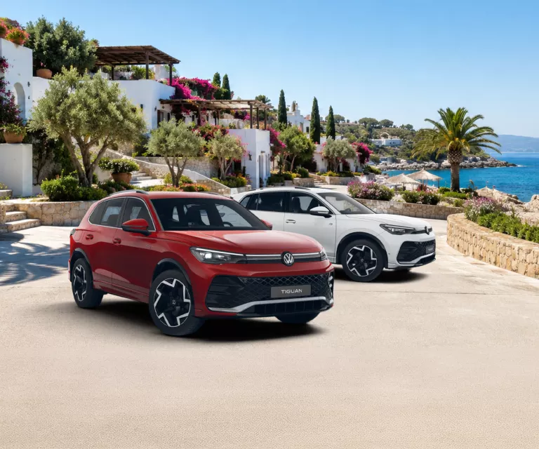 Zwei VW Tiguan, einer rot und einer weiß, stehen vor mediterraner Küstenlandschaft mit Meer und weißen Häusern.