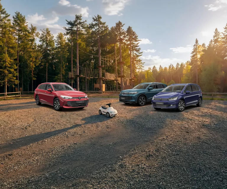 Drei VW Modelle stehen bei untergehnder Sonne vor einem Kletterpark auf einem Parkplatz im Wald.