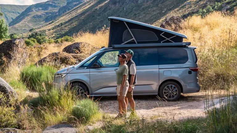 Silberner Volkswagen Nutzfahrzeug-Camper mit aufgeklapptem Dach in einer bergigen Landschaft, zwei Personen stehen davor und schauen nach oben