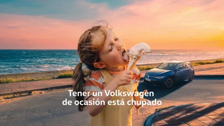 Una niña disfrutando de un cucurucho con un Volkswagen azul detrás en la playa