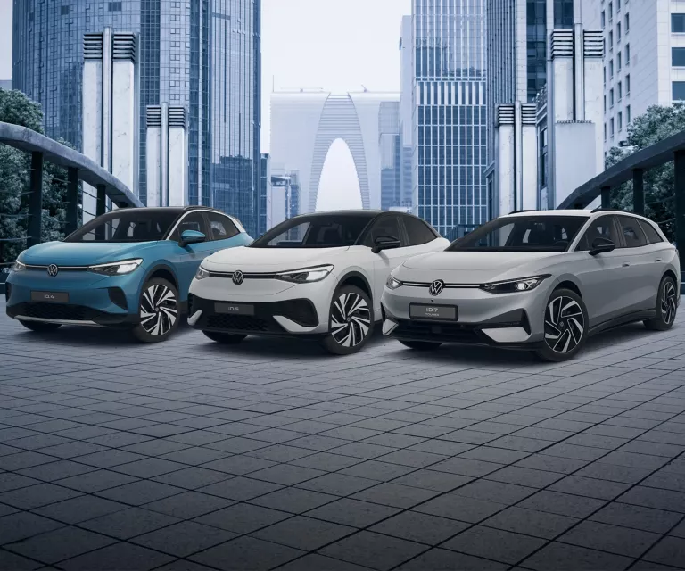 Ein blauer VW ID.4, ein weißer VW ID.5 und ein grauer VW ID.7 Tourer stehen auf einer gepflasterten Fläche in einer Stadt.