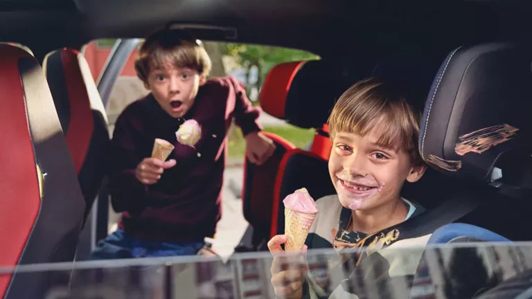 Zwei Kinder sitzen mit einem Eis in einem Auto. Einem Kind fällt die Kugel Eis aus der Waffel auf den Sitz.